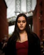 A woman with long dark hair wearing a maroon top and a black coat is standing outdoors in an urban environment. The background features a blurred view of a bridge and two brick buildings on either side.