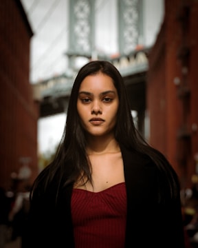 A woman with long dark hair wearing a maroon top and a black coat is standing outdoors in an urban environment. The background features a blurred view of a bridge and two brick buildings on either side.