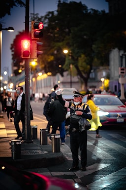 A traffic officer in uniform is standing on a city street during evening time. The traffic light above is red, and several pedestrians are walking nearby. A car is seen in the background, while the officer holds up a hand to manage the traffic flow. The atmosphere appears bustling with city activity.