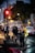 A traffic officer in uniform is standing on a city street during evening time. The traffic light above is red, and several pedestrians are walking nearby. A car is seen in the background, while the officer holds up a hand to manage the traffic flow. The atmosphere appears bustling with city activity.