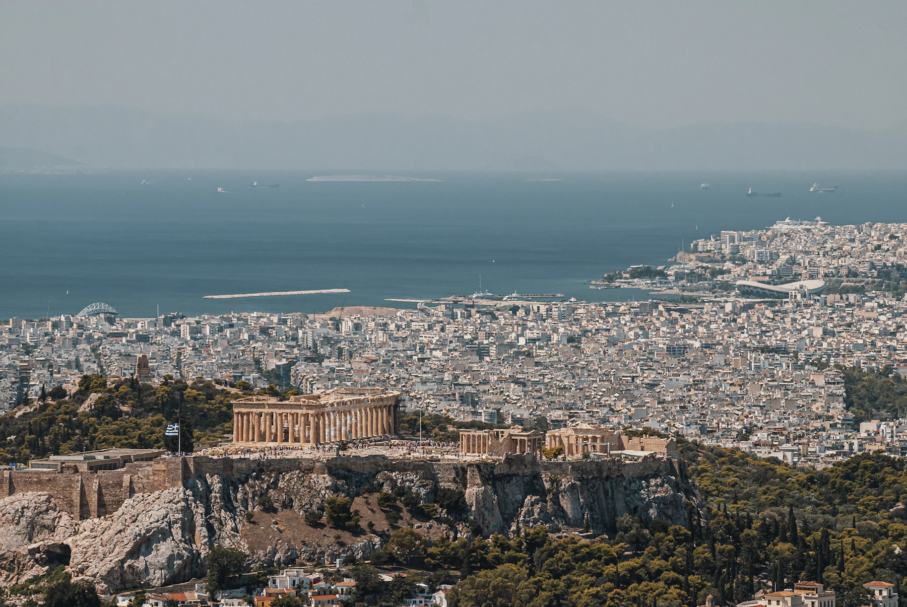 Athens seen whole from above, the Acropolis small against the city, the Saronic Gulf beyond.