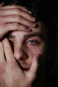 A close-up of a person’s face with hands covering parts of their face. Blue eyes with a tear, rough stubble, and expressive features suggest emotion.