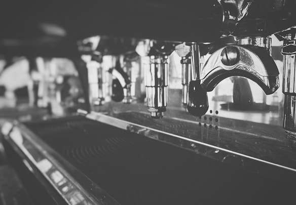 A close-up view of a stainless steel espresso machine, showcasing shiny metallic components including a portafilter and various levers and pipes. The image is in black and white, highlighting the sleek and polished surfaces with reflections.
