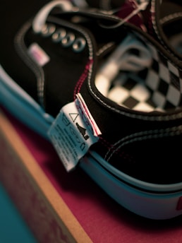 Close-up view of a black sneaker with white stitching and metal eyelets, displaying a checkered pattern on the interior. A textile care label is attached to the shoe, and it is placed on a pink and brown box.