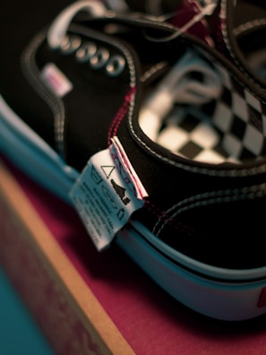 Close-up view of a black sneaker with white stitching and metal eyelets, displaying a checkered pattern on the interior. A textile care label is attached to the shoe, and it is placed on a pink and brown box.