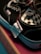 Close-up view of a black sneaker with white stitching and metal eyelets, displaying a checkered pattern on the interior. A textile care label is attached to the shoe, and it is placed on a pink and brown box.