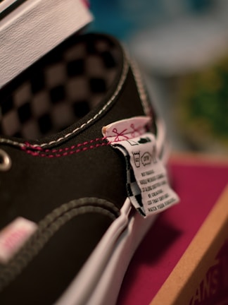 A close-up view of a black shoe with white stitching and a checkered inner lining. Partially visible, a label with washing instructions and small icon graphics is attached near the tongue area. The shoe appears to be placed on a surface with a maroon and brown box nearby.