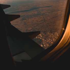 An aerial view of a landscape, with a city partially visible through the airplane window. The image is illuminated by a warm, golden light suggesting either sunrise or sunset.