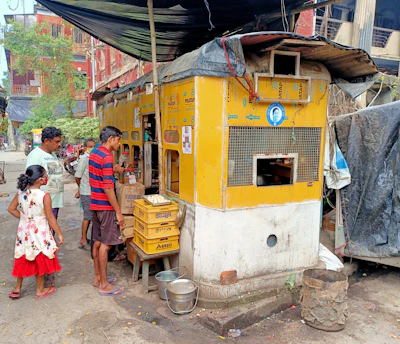 A small street-side shop with a yellow painted exterior and corrugated metal roof is shown. Several people, including a man and a child in a red dress, are standing nearby. The shop appears to be selling or distributing some goods with stacks of crates and utility items like metal buckets visible outside.