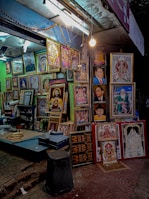 A street-side shop displays a variety of framed portraits and religious artwork. The shop is well-lit with a hanging bulb and fluorescent lights. The frames are arranged closely together, covering the wall and standing on the floor, showcasing different styles and colors. The setting is vibrant and colorful, providing a glimpse into a busy street market ambiance.