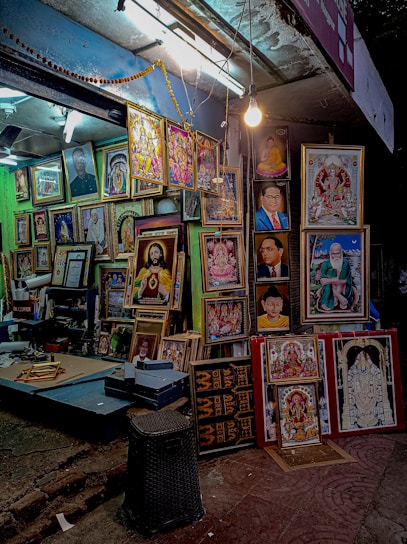 A street-side shop displays a variety of framed portraits and religious artwork. The shop is well-lit with a hanging bulb and fluorescent lights. The frames are arranged closely together, covering the wall and standing on the floor, showcasing different styles and colors. The setting is vibrant and colorful, providing a glimpse into a busy street market ambiance.