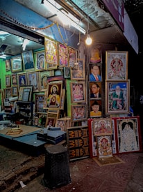A street-side shop displays a variety of framed portraits and religious artwork. The shop is well-lit with a hanging bulb and fluorescent lights. The frames are arranged closely together, covering the wall and standing on the floor, showcasing different styles and colors. The setting is vibrant and colorful, providing a glimpse into a busy street market ambiance.