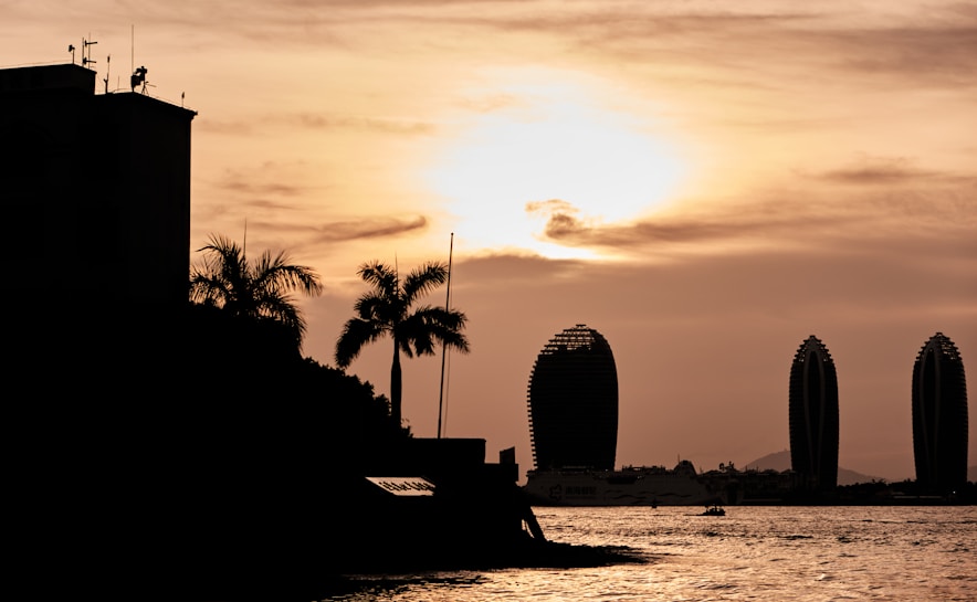 The image captures a silhouette of palm trees and modern buildings against a sunset sky. The skyline features uniquely shaped, tall buildings, and the sun casts a warm glow over the water, creating a serene and picturesque scene.