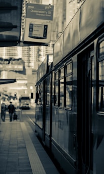 A tram is parked at a city street tram stop, under a sign indicating the location as Queensberry Street on Swanston Street. Buildings are visible in the background, along with pedestrians walking by and cars on the street.