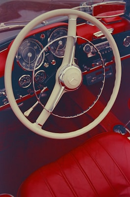 A vintage car interior with a glossy red leather seat and dashboard. The classic steering wheel is white and chrome, and there are multiple gauges on the dashboard. The design is elegant and retro, evoking a sense of nostalgia.