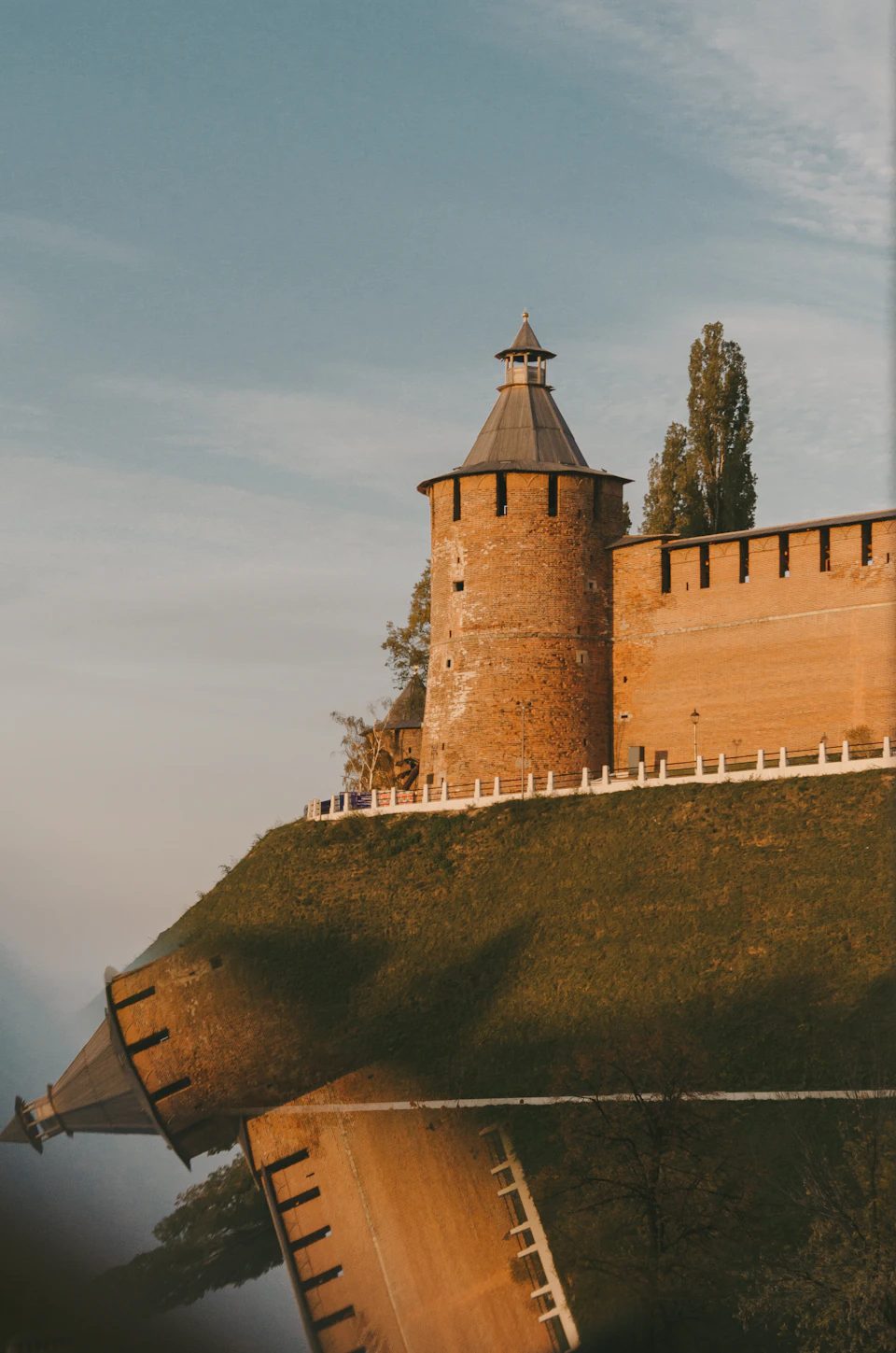 The image depicts a large, orange brick tower with a pointed roof and a small tower on top, situated atop a hill. The tower is surrounded by lush green trees and grass, creating a picturesque scene. In the foreground, there are two people walking along the path leading up to the tower.