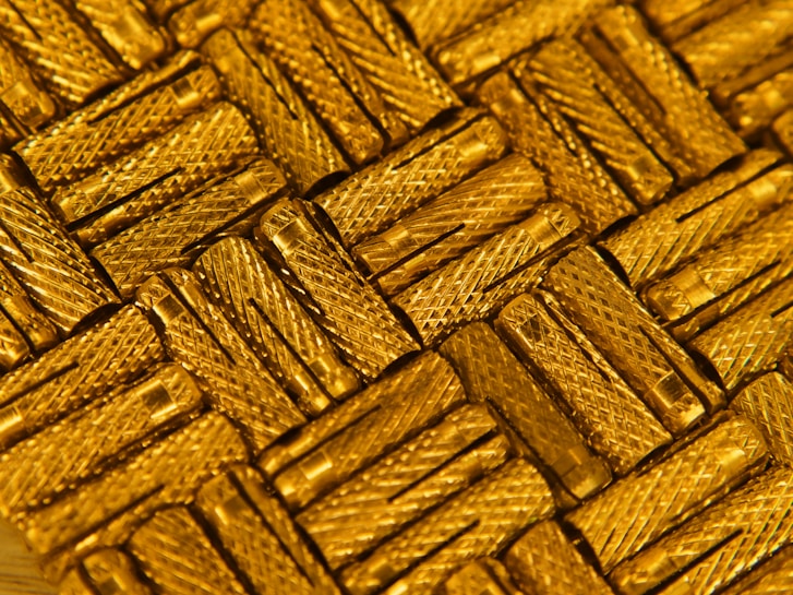 A close-up view of intricately arranged gold-colored metal pieces with a textured, crisscross pattern. The light reflects off the metallic surface, enhancing the luxurious appearance.