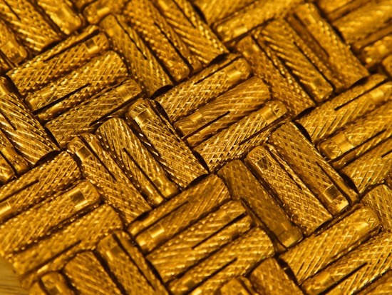 A close-up view of intricately arranged gold-colored metal pieces with a textured, crisscross pattern. The light reflects off the metallic surface, enhancing the luxurious appearance.