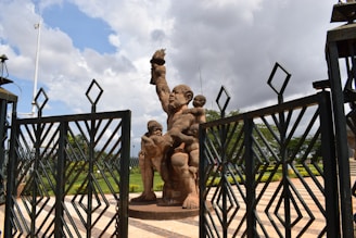 A monumental statue depicting a large figure holding a flame with smaller figures around its legs. The scene is framed by large metal gates, and the area appears to be part of a public square or monument site. Greenery and patterned stone paving enhance the background, while clouds scatter across the sky.