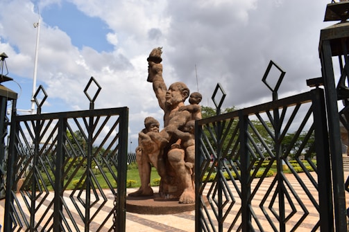 A monumental statue depicting a large figure holding a flame with smaller figures around its legs. The scene is framed by large metal gates, and the area appears to be part of a public square or monument site. Greenery and patterned stone paving enhance the background, while clouds scatter across the sky.
