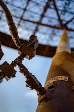 A close-up view of a rusty mechanical structure featuring interlocking, weathered gears and metal components. The background reveals an industrial framework against a blue sky with sparse clouds.