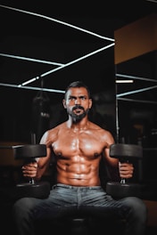 A muscular man is seated on a bench, holding dumbbells in each hand. He has a well-defined six-pack and is in a gym setting. The background features modern, geometric ceiling lights and a dark, industrial atmosphere.