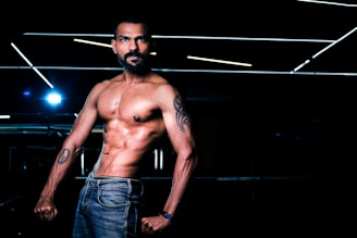 A muscular man with tattoos poses shirtless, displaying his toned physique. His jeans add a casual element, while the dark background is punctuated with horizontal fluorescent lighting, creating a dramatic and focused atmosphere.