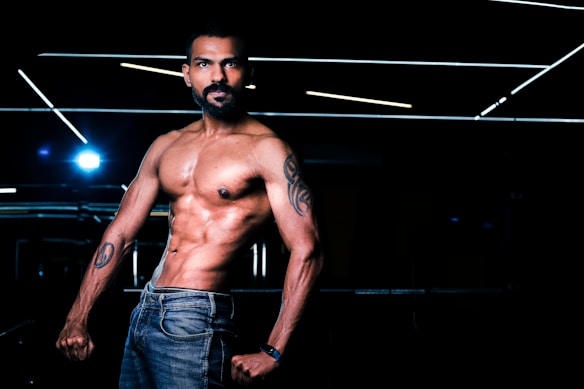 A muscular man with tattoos poses shirtless, displaying his toned physique. His jeans add a casual element, while the dark background is punctuated with horizontal fluorescent lighting, creating a dramatic and focused atmosphere.