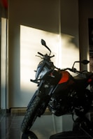 A motorcycle with a sharp and rugged design is positioned inside a room with sunlight casting shadows on the wall. The bike has a striking combination of black and orange colors, large tires, and a sturdy build, suggesting it is designed for on-road and off-road use.