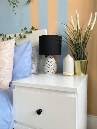 A neatly arranged bedside table features a white drawer with a black knob, topped with a speckled lamp, a minimalist white vase, and a potted plant. The background showcases a striped wall with blue and beige tones, while pillows with light blue and patterned covers are visible on the bed.