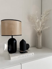 A minimalist interior setting features a lamp with a woven rattan shade and black base, a black diffuser, a small black speaker, and a book titled 'Live Beautiful' on a white cabinet. A vase with dried, white leaves adds a natural element to the composition.