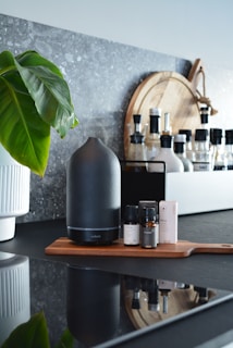 A modern kitchen counter features a sleek black aroma diffuser placed on a wooden tray alongside several small bottles and a box, suggesting essential oils. A large green leaf plant in a white pot adds a touch of nature. In the background, various condiment bottles and a wooden cutting board provide a homely, organized atmosphere.