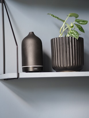A minimalist shelf features a dark ceramic essential oil diffuser next to a modern, ribbed planter with a green plant. The backdrop is a soft blue wall, contributing to a calming atmosphere.