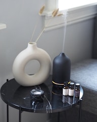 A round black marble table with a white vase holding dried grass stems, a black diffuser emitting mist, four small essential oil bottles, and a small marble container.