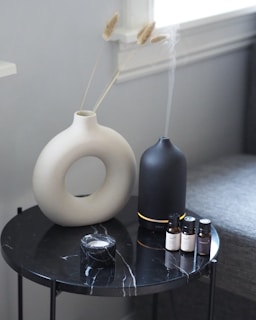 A round black marble table with a white vase holding dried grass stems, a black diffuser emitting mist, four small essential oil bottles, and a small marble container.