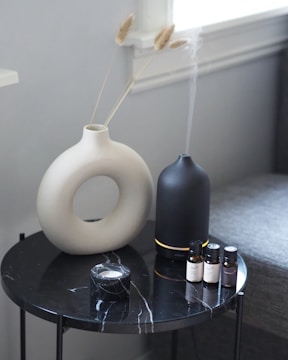 A round black marble table with a white vase holding dried grass stems, a black diffuser emitting mist, four small essential oil bottles, and a small marble container.