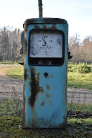 An old, rusty gas pump situated in an outdoor setting. The pump is mostly blue with significant rust patches covering its surface. It features an analog display dial, showing numbers and a needle. The background consists of a natural landscape with grass, trees, and a dirt path.