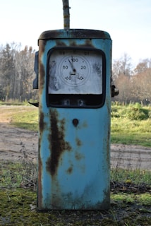An old, rusty gas pump situated in an outdoor setting. The pump is mostly blue with significant rust patches covering its surface. It features an analog display dial, showing numbers and a needle. The background consists of a natural landscape with grass, trees, and a dirt path.