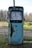 An old, rusty gas pump situated in an outdoor setting. The pump is mostly blue with significant rust patches covering its surface. It features an analog display dial, showing numbers and a needle. The background consists of a natural landscape with grass, trees, and a dirt path.