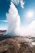 A smiling woman standing near Old Faithful geyser with steam rising in the background under a clear blue sky.