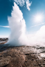A large geyser erupts, shooting water and steam high into the air against a clear blue sky. The bright sun shines, casting light over the rocky ground. A group of people stands at a distance, observing the natural phenomenon.