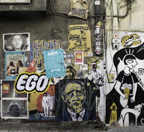 A wall covered with a variety of street art, including posters, stickers, and graffiti. Among the artwork are colorful illustrations, abstract faces, and text, such as the word 'EGO' in large letters. The styles range from cartoonish black-and-white drawings to vibrant, expressive portraits. Some images include human figures and surreal elements, contributing to a vibrant and eclectic visual tapestry.