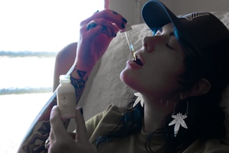 A person wearing a cap and dangling earrings shaped like leaves is using a dropper to take a liquid from a small bottle. Their arm has visible tattoos, and they are seated in a softly lit room with a window, suggesting natural light.