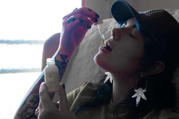 A person wearing a cap and dangling earrings shaped like leaves is using a dropper to take a liquid from a small bottle. Their arm has visible tattoos, and they are seated in a softly lit room with a window, suggesting natural light.