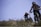 Two mountain bikers wearing protective gear and helmets are riding down a rocky and grassy trail. The scene is set outdoors with clear blue skies and natural greenery, indicating a bright and adventurous atmosphere. The focus is on the bikers who appear to be engaged in a downhill ride.