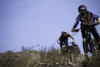 Two mountain bikers wearing protective gear and helmets are riding down a rocky and grassy trail. The scene is set outdoors with clear blue skies and natural greenery, indicating a bright and adventurous atmosphere. The focus is on the bikers who appear to be engaged in a downhill ride.