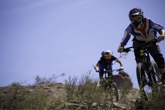 Two mountain bikers wearing protective gear and helmets are riding down a rocky and grassy trail. The scene is set outdoors with clear blue skies and natural greenery, indicating a bright and adventurous atmosphere. The focus is on the bikers who appear to be engaged in a downhill ride.