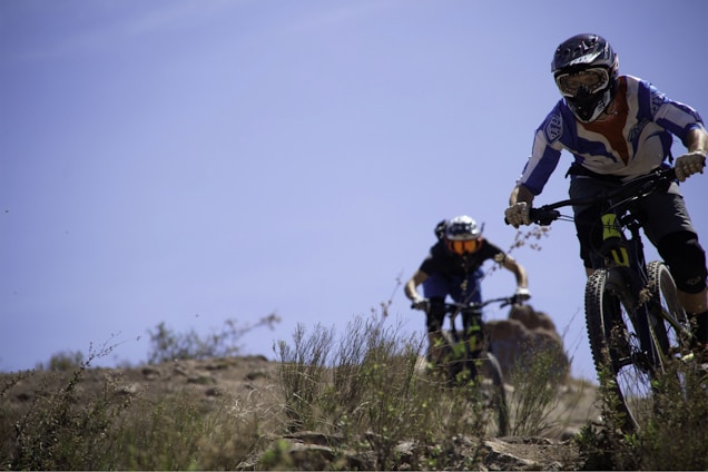 Two mountain bikers wearing protective gear and helmets are riding down a rocky and grassy trail. The scene is set outdoors with clear blue skies and natural greenery, indicating a bright and adventurous atmosphere. The focus is on the bikers who appear to be engaged in a downhill ride.