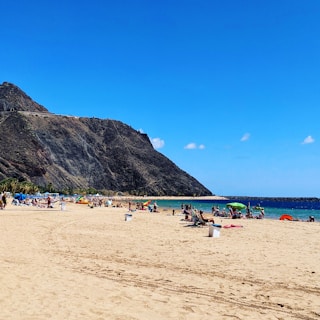 A sandy beach with people relaxing under umbrellas, set against a backdrop of rocky cliffs and a clear blue sky. The sea is calm with small waves and vibrant turquoise hues.