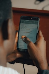 A person is holding a smartphone with both hands, reading a chat conversation on the screen. The chat messages are visible, containing text and emojis. The background is blurred and has a warm tone, indicating a possibly indoor environment.